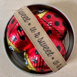 May include: A silver tin with a brown ribbon that says "so sweet - u r so sweet - u r so" and three red and black ladybug-shaped chocolates inside.
