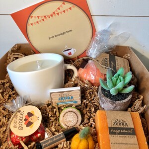 May include: A gift box filled with fall-themed items. Includes a white mug candle, pumpkin-shaped bath bomb, succulent, orange oatmeal soap, and a card that says "your message here". The box is filled with brown paper and has a festive autumn theme.