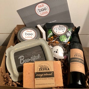 May include: A Halloween-themed gift box filled with various items, including a framed sign that says "You are Boo-tiful", a jar of candle with a label that says "FAB {boo} LOUS", a bar of orange oatmeal handmade soap, a bottle of cinnamon pumpkin luxurious lotion, a bag of chocolate candies with a label that says "YUMMY boo", and a white ghost plush toy.