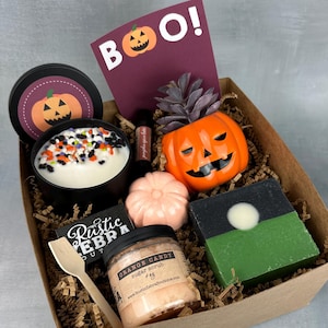 May include: A brown cardboard box filled with Halloween themed bath and body products. The box contains a pumpkin-shaped candle, a pumpkin-shaped soap, a pumpkin-shaped sugar scrub, a pumpkin-shaped soap dish, and a small plant in a pumpkin-shaped pot. The box also includes a wooden spoon and a card that says "BOO!"
