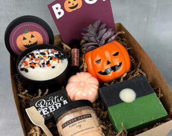 May include: A brown cardboard box filled with Halloween themed bath and body products. The box contains a pumpkin-shaped candle, a pumpkin-shaped soap, a pumpkin-shaped sugar scrub, a pumpkin-shaped soap dish, and a small plant in a pumpkin-shaped pot. The box also includes a wooden spoon and a card that says "BOO!"