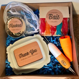 Puede incluir: Una caja de regalo llena de art&iacute;culos con tem&aacute;tica de pesca, que incluyen una red, un peque&ntilde;o marco de fotos con el texto "Gracias - Por todo lo que haces", un flotador, un se&ntilde;uelo de pesca y una bolsa de peces de caramelo con el texto "MMM Bait YUM".