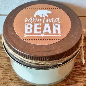 May include: A brown jar candle with a tan lid and twine wrapped around the lid. The lid has a round, brown label with a white bear silhouette, an arrow, a heart, and the text "momma BEAR".