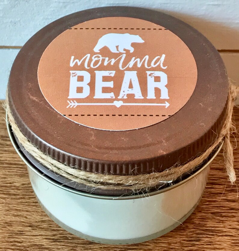 May include: A brown jar candle with a tan lid and twine wrapped around the lid. The lid has a round, brown label with a white bear silhouette, an arrow, a heart, and the text "momma BEAR".