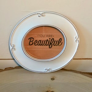 May include: White oval frame with a distressed finish and a brown background with the text "You are Beautiful" in black lettering.