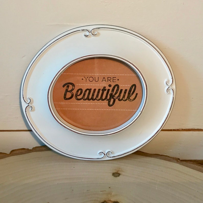 May include: White oval frame with a distressed finish and a brown background with the text "You are Beautiful" in black lettering.