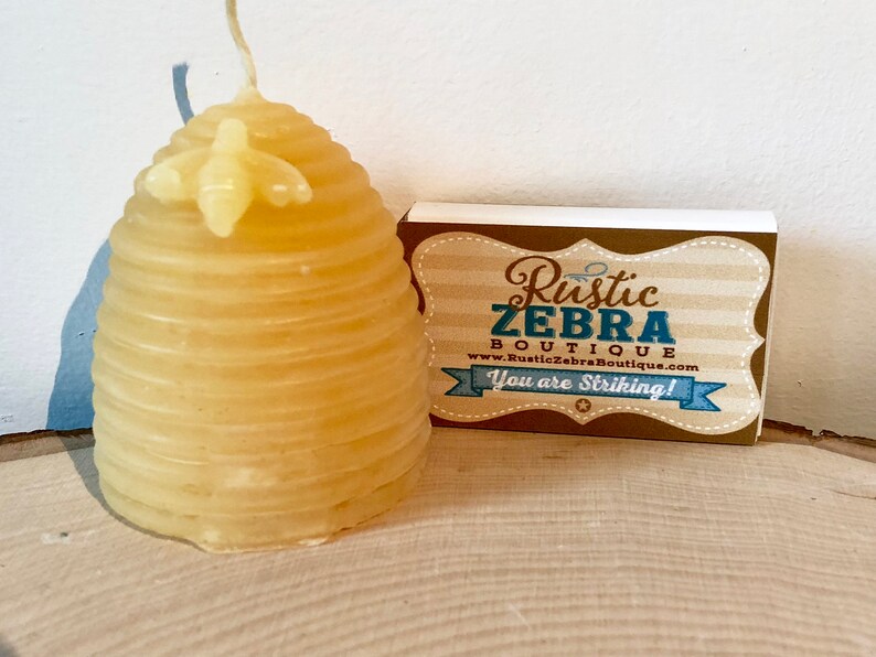May include: A yellow beeswax candle shaped like a beehive with a small bee on top. The candle is sitting on a wooden surface next to a business card that reads "Rustic Zebra Boutique" and "You are Striking!"