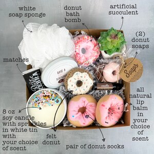 May include: A gift box filled with donut-themed items, including a bath bomb, soap, socks, a candle, and a lip balm. The box also includes a white soap sponge, matches, and an artificial succulent.