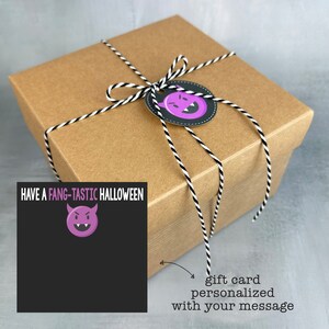 May include: A brown gift box tied with black and white twine, featuring a Halloween-themed tag with a purple devil face. A black card with the text "HAVE A FANG-TASTIC HALLOWEEN" and a purple devil graphic is attached.