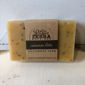 Puede incluir: Una barra de jab&oacute;n artesanal de canela y latte con una etiqueta de papel marr&oacute;n que dice "Rustic Zebra Boutique" y "jab&oacute;n artesanal de canela y latte", junto con la direcci&oacute;n del sitio web "www.RusticZebraBoutique.com".