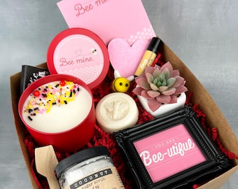 May include: A gift box filled with bee-themed items, including a candle, soap, sugar scrub, and a small succulent plant. The box is decorated with red and white tissue paper and a pink card that says "Bee mine."