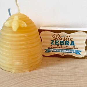 May include: A yellow beeswax candle shaped like a beehive with a small bee on top. The candle is sitting on a wooden surface next to a business card that reads "Rustic Zebra Boutique" and "You are Striking!"