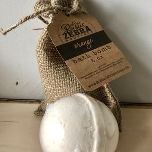 May include: A white bath bomb, 5 oz, sits in front of a burlap bag with a tag that reads "Rustic Zebra Boutique, orange, bath bomb." The bag is tied with twine. The bath bomb is wrapped in clear plastic.