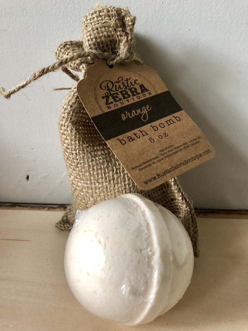 May include: A white bath bomb, 5 oz, sits in front of a burlap bag with a tag that reads "Rustic Zebra Boutique, orange, bath bomb." The bag is tied with twine. The bath bomb is wrapped in clear plastic.