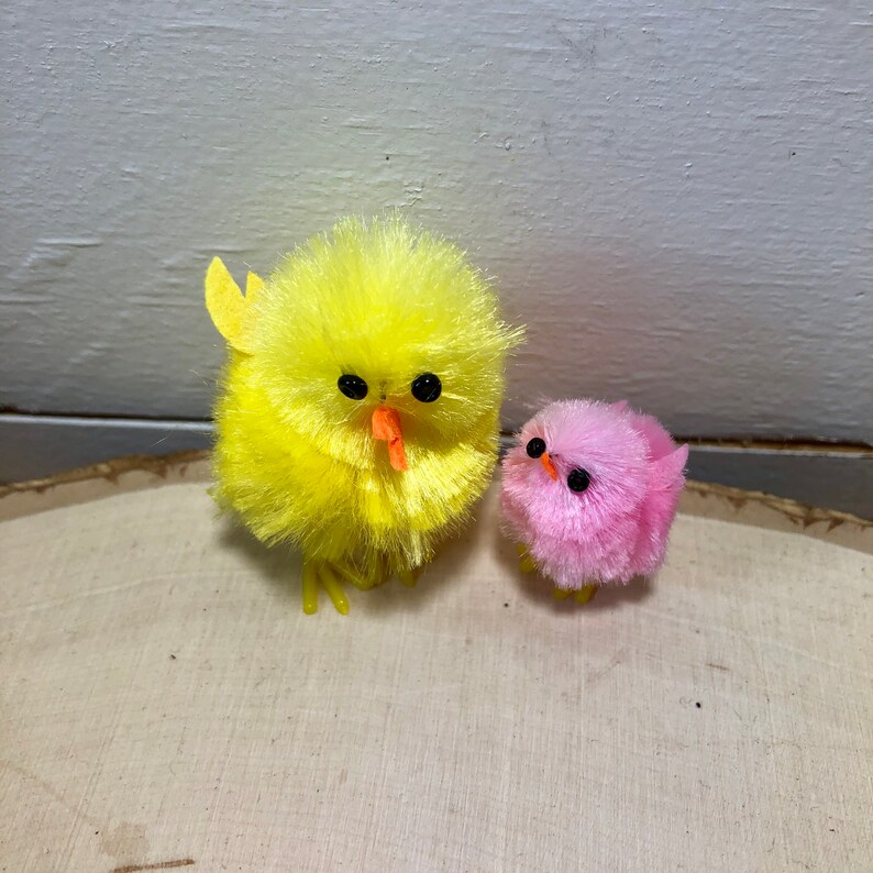 May include: Two fluffy chicks, one yellow and one pink, with black eyes and orange beaks. They are both standing on a wooden surface.