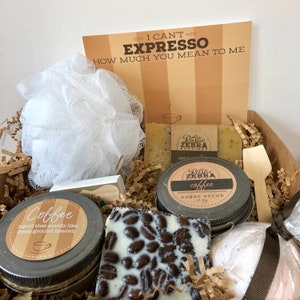 May include: A gift box filled with coffee-themed bath and body products. The box includes a coffee-scented sugar scrub, a coffee-scented bath bomb, a coffee-scented soap, and a coffee-scented body wash. The box also includes a small wooden spoon and a white washcloth. The box is decorated with brown paper shreds and a card that reads "I can't espresso how much you mean to me."