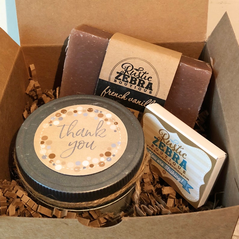 K&ouml;nnte beinhalten: Eine Geschenkbox gef&uuml;llt mit braunem Seidenpapier, die eine Kerze mit einem "Danke"-Etikett, eine Seife mit einem "franz&ouml;sisches Vanille"-Etikett und ein kleines Holzschild mit dem Text "Rustic Zebra Boutique You are Striking!" enth&auml;lt.