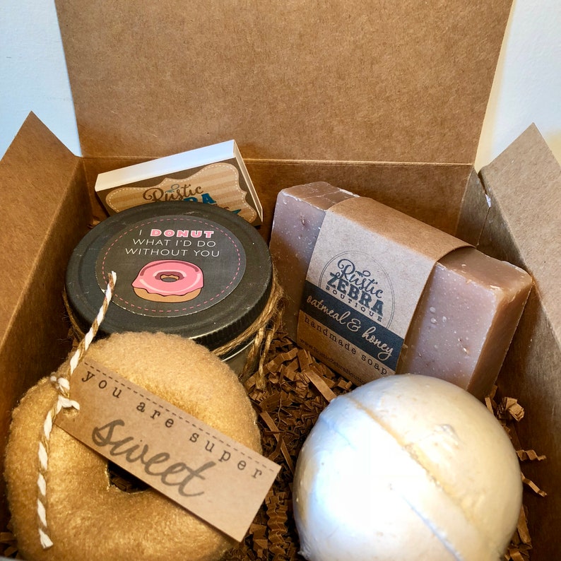 May include: A gift box filled with bath and body products. The box contains a jar of bath scrub with a donut-themed label that says "I donut what I'd do without you", a bar of oatmeal and honey soap with a Rustic Zebra Boutique label, a bath bomb, and a donut-shaped plush toy with a tag that says "You are super sweet".