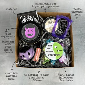 May include: A Halloween-themed gift box containing a 4 oz soy candle in a black tin, a small lotion bar, plastic vampire teeth, a small felt vampire head, lip balm, matches, and a bag of chocolates. The candle has a purple devil face on the lid.
