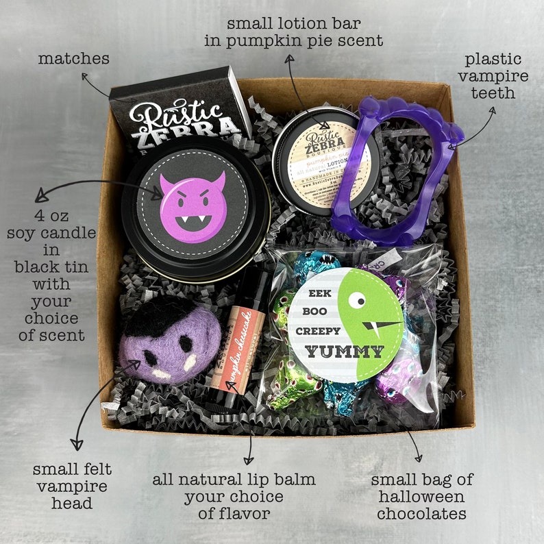May include: A Halloween-themed gift box containing a 4 oz soy candle in a black tin, a small lotion bar, plastic vampire teeth, a small felt vampire head, lip balm, matches, and a bag of chocolates. The candle has a purple devil face on the lid.