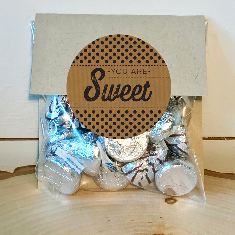 Op de afbeelding: Een bruine papieren zak met een ronde sticker waarop in zwarte tekst "You are Sweet" staat op een bruine stippenachtergrond. De zak bevat een doorzichtige plastic zak gevuld met zilveren, in folie verpakte chocoladekusjes.