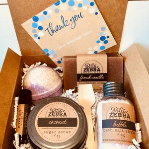 May include: A gift box filled with bath and body products. The box contains a bath bomb, a sugar scrub, bath salts, a bar of soap, and a lip balm. The products are all from the Rustic Zebra Boutique.