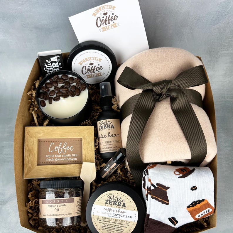 May include: A brown gift box filled with coffee-themed items, including a candle, lotion bar, sugar scrub, socks, and a throw blanket. The box is decorated with brown paper and a brown ribbon. The text on the items reads "It's Always Coffee Time", "Rustic Zebra Boutique", "Coffee", and "Coffee Shop Lotion Bar".