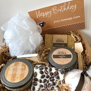 May include: A gift basket filled with coffee-themed bath and body products. The basket includes a white bath pouf, a bar of cinnamon latte soap, a jar of coffee sugar scrub, a bar of white chocolate with coffee beans, and a bath bomb. The basket is decorated with brown paper shreds and a card that says "Happy Birthday" with space for a personalized message.