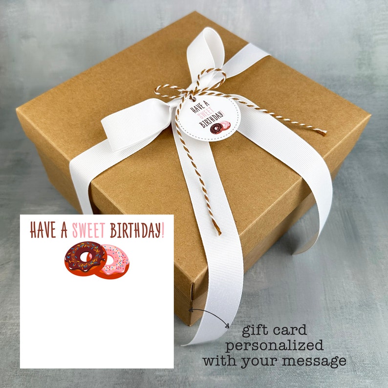 May include: A brown gift box with a white ribbon and a gift tag that says "Have a Sweet Birthday!" The gift tag has a pink and brown donut illustration. A white card with the same text and donut illustration is in the foreground. The card has space for a personalised message.