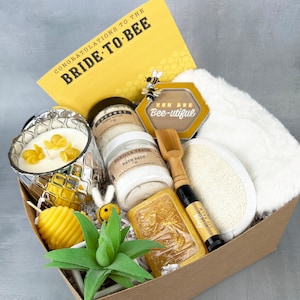 May include: A brown gift box filled with a variety of items, including a candle, bath salts, soap, lip balm, a wooden spoon, a white washcloth, and a small green plant. The box is decorated with a yellow card that reads "Congratulations to the Bride-to-Bee" and a yellow hexagonal tag that reads "You are Bee-utiful".