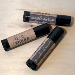 May include: Three black lip balm tubes with brown paper labels. The labels have the text "Rustic Zebra Boutique" and the flavors "maple syrup", "pumpkin cheesecake", and "all natural lip balm".