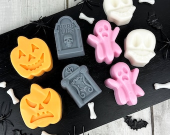 May include: A collection of Halloween-themed wax melts in various shapes, including jack-o'-lanterns, tombstones, ghosts, skulls, and bones. The melts are orange, gray, pink, and white, arranged on a black surface with decorative spiders and bats.