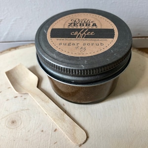 Puede incluir: Un peque&ntilde;o frasco de vidrio con tapa negra y una etiqueta marr&oacute;n que dice "Rustic Zebra Boutique coffee sugar scrub 4 oz" y "www.RusticZebraBoutique.com". El frasco est&aacute; sobre una superficie de madera con una peque&ntilde;a cuchara de madera al lado.