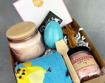 May include: A brown cardboard gift box filled with Easter-themed items, including a candle, a blue egg, a wooden cross, a pair of socks with a yellow chick design, and a jar of strawberry sugar scrub. The box is decorated with shredded paper and a card that says "Happy Easter".