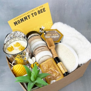 May include: A gift basket filled with bath and body products, including a candle, bath salts, soap, lip balm, and a loofah. The basket is decorated with a yellow and black bee theme and includes a card that says "Congratulations to the Mommy-to-Bee".