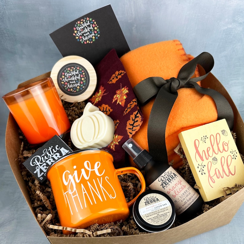 May include: An autumn-themed gift box filled with an orange mug that says "give thanks", a candle, a small blanket, a bar of soap shaped like a pumpkin, and other fall-themed items. The box includes a card that says "hello fall".