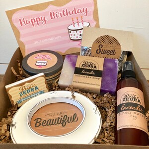 May include: A gift box with a birthday theme. It contains a card that says "Happy Birthday", a candle, soap, a picture frame with the words "You are Beautiful", and a lavender linen spray. The box is filled with brown paper.