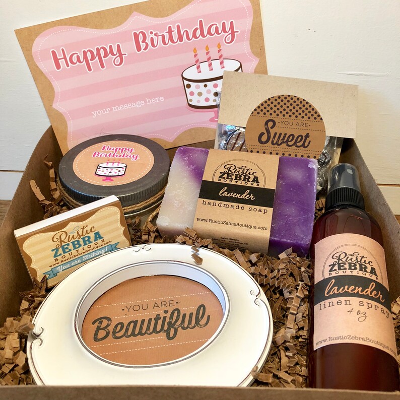 May include: A gift box with a birthday theme. It contains a card that says "Happy Birthday", a candle, soap, a picture frame with the words "You are Beautiful", and a lavender linen spray. The box is filled with brown paper.