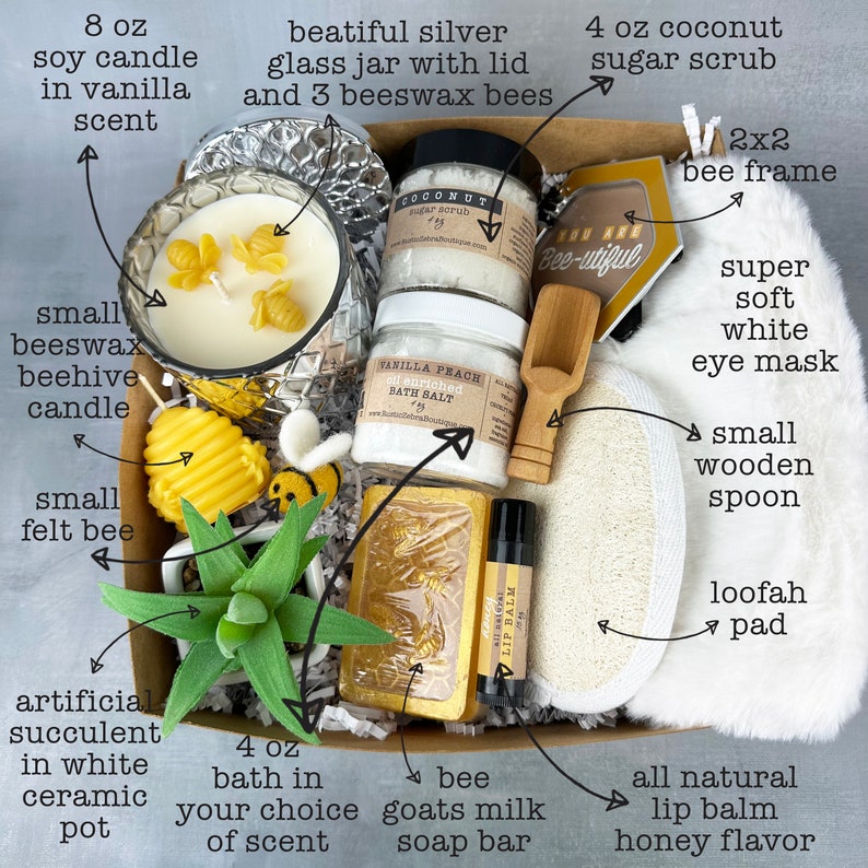 May include: A gift box filled with bath and body products, including a 4 oz. coconut sugar scrub, a 4 oz. bath salt, a 4 oz. goat milk soap bar, a 8 oz. soy candle, a small beeswax beehive candle, a small wooden spoon, a loofah pad, a white eye mask, a lip balm, a felt bee, a succulent in a white ceramic pot, and a 2x2 bee frame. The box is decorated with a bee-themed design.