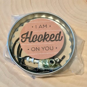 Puede incluir: Una peque&ntilde;a lata redonda con un se&ntilde;uelo de pesca en su interior. La lata tiene una etiqueta de papel marr&oacute;n con el texto "I am Hooked on You".