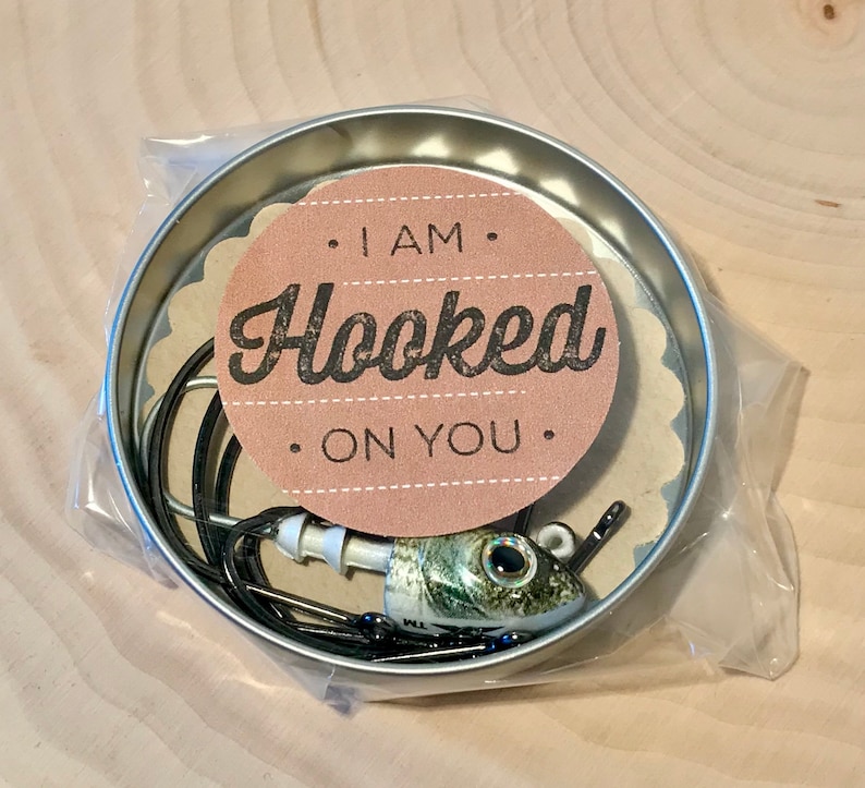 Puede incluir: Una peque&ntilde;a lata redonda con un se&ntilde;uelo de pesca en su interior. La lata tiene una etiqueta de papel marr&oacute;n con el texto "I am Hooked on You".