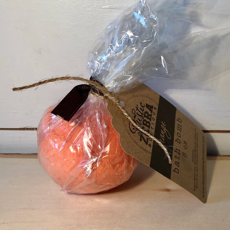 May include: An orange bath bomb, wrapped in clear cellophane and tied with twine and a brown ribbon. A brown tag with the text "Rustic Zebra Boutique" and "Orange bath bomb 5 oz" is attached.