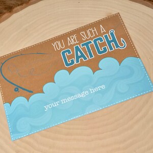 Puede incluir: Una tarjeta marr&oacute;n con un dise&ntilde;o de olas azul y blanco. La tarjeta dice "You are such a CATCH" con un anzuelo. Hay una ca&ntilde;a de pescar y una silueta de pez en el fondo. La tarjeta tiene un espacio para un mensaje.