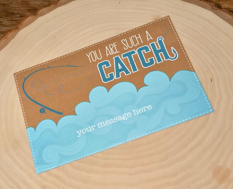 Puede incluir: Una tarjeta marr&oacute;n con un dise&ntilde;o de olas azul y blanco. La tarjeta dice "You are such a CATCH" con un anzuelo. Hay una ca&ntilde;a de pescar y una silueta de pez en el fondo. La tarjeta tiene un espacio para un mensaje.