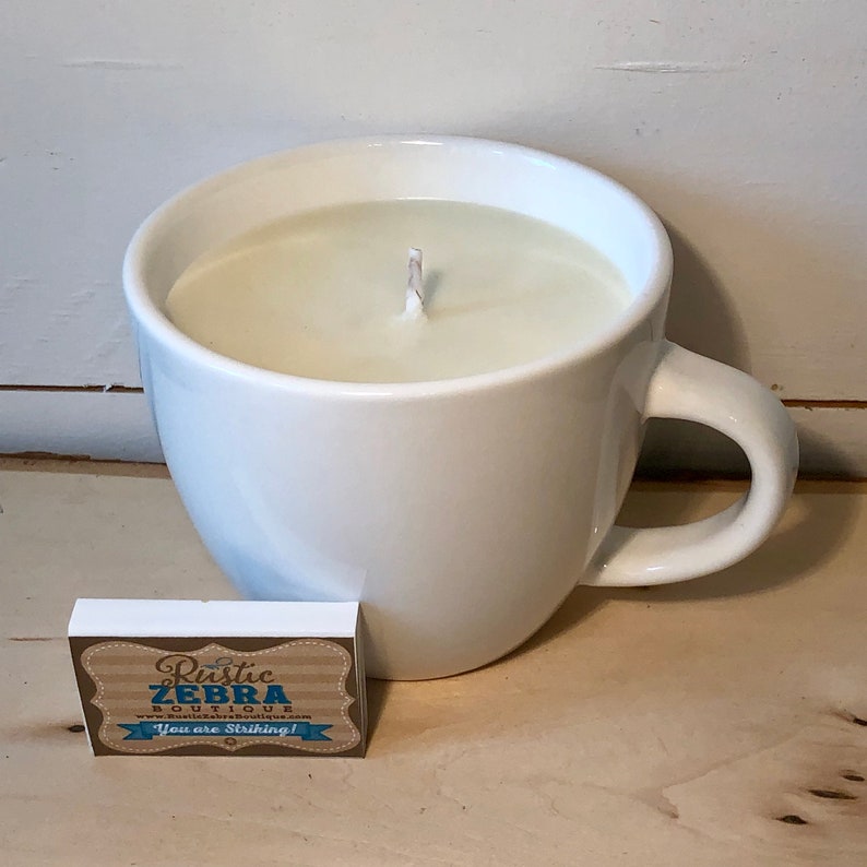 May include: A white ceramic mug filled with a cream-colored candle. A matchbox with the text "Rustic Zebra Boutique" and "You are Striking!" is next to the mug. The candle has a single wick.