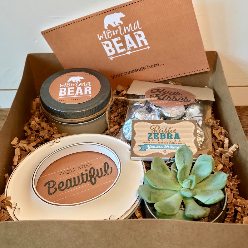 May include: A gift box filled with brown shredded paper, a candle with a "momma bear" label, a small succulent plant in a pot, a small plate with a "you are beautiful" label, and a bag of chocolate kisses with a "hugs kisses" label.
