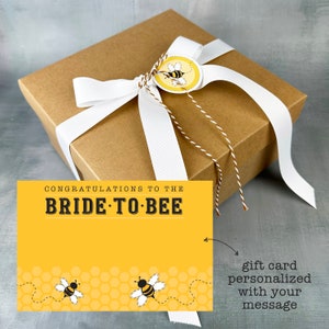 May include: A brown gift box tied with a white ribbon and a bee-themed gift tag. The box is sitting on a gray surface. A yellow card with the text "Congratulations to the Bride-to-Bee" is in front of the box. The card has a bee illustration and honeycomb pattern. A handwritten note says "gift card personalized with your message".