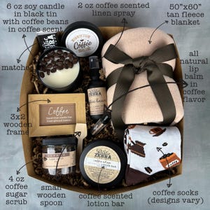 May include: A coffee-themed gift basket with a tan fleece blanket, coffee-scented lotion bar, coffee socks, a small wooden spoon, coffee sugar scrub, a 3x2 wooden frame with the word "Coffee" on it, a 6 oz soy candle in a black tin with coffee beans, a 2 oz coffee-scented linen spray, and a lip balm in coffee flavor.