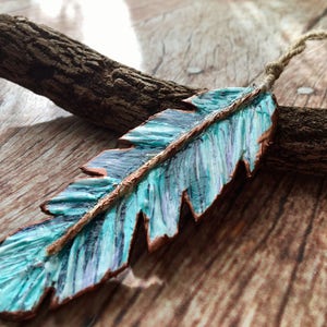 May include: A teal blue and copper feather ornament with a rustic twine hanger. The feather is painted with a marbled effect and has a copper wire running down the center.