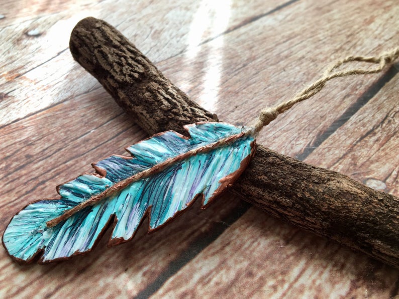 May include: A turquoise blue and copper colored feather ornament with a brown twine hanger. The feather is painted with a striped pattern and has a rustic look.